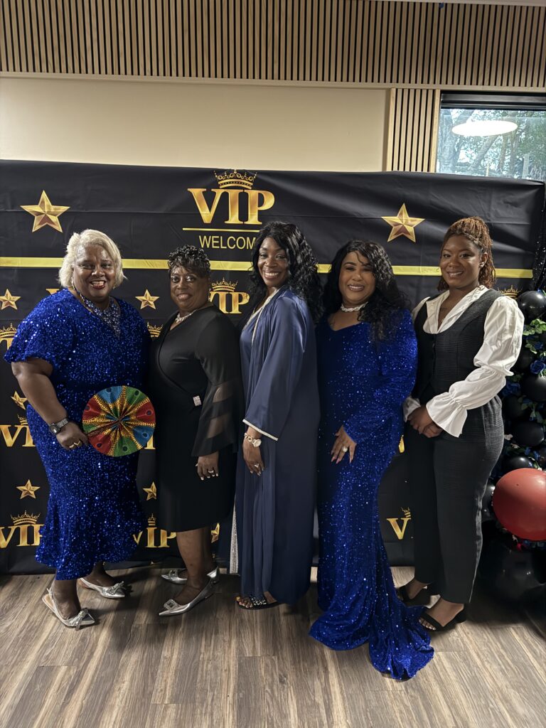 Five women in formal attire posing at a VIP event photo backdrop.