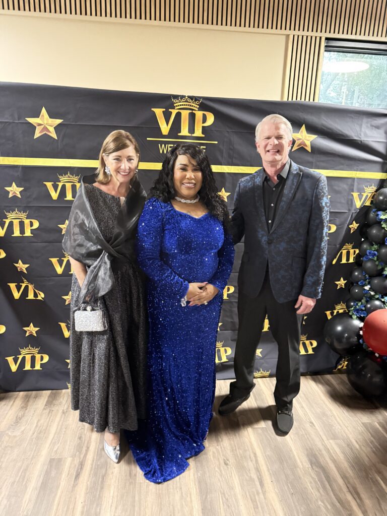 Three people dressed elegantly at a VIP event with a gold and black backdrop.