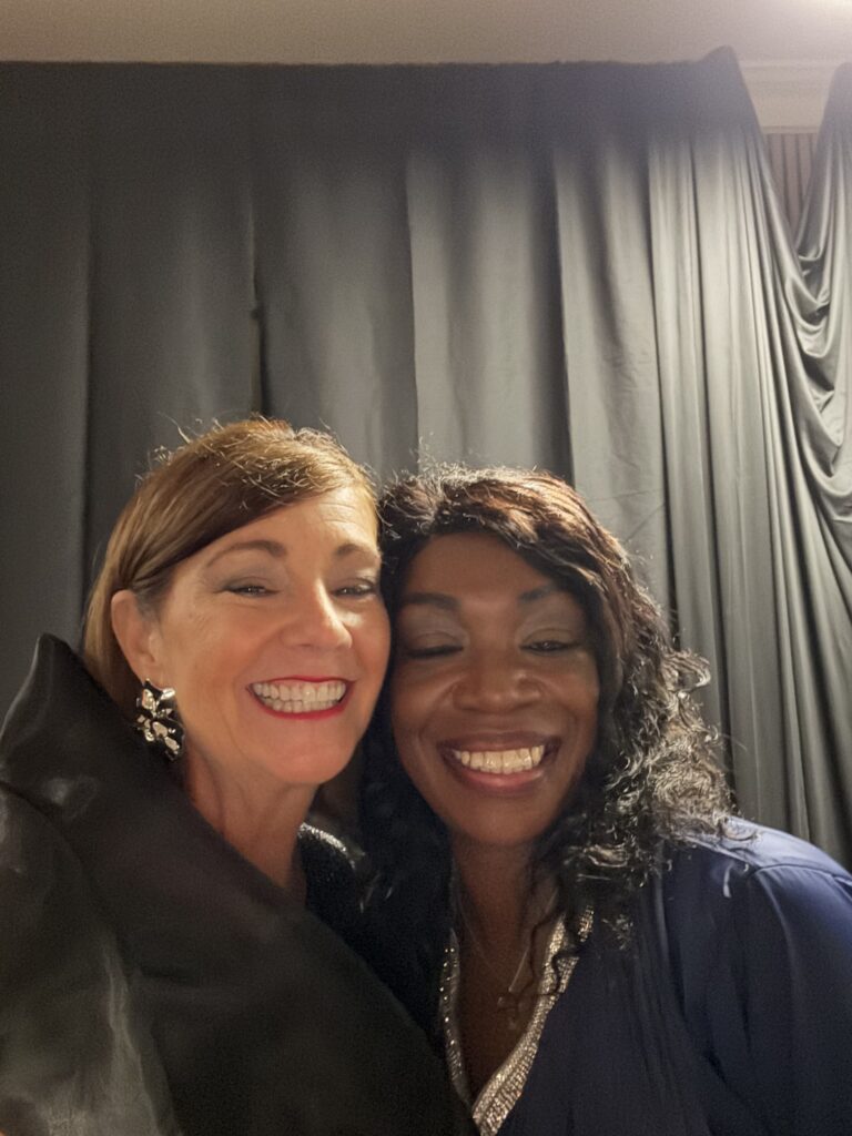 Two women smiling warmly in front of a dark curtain.